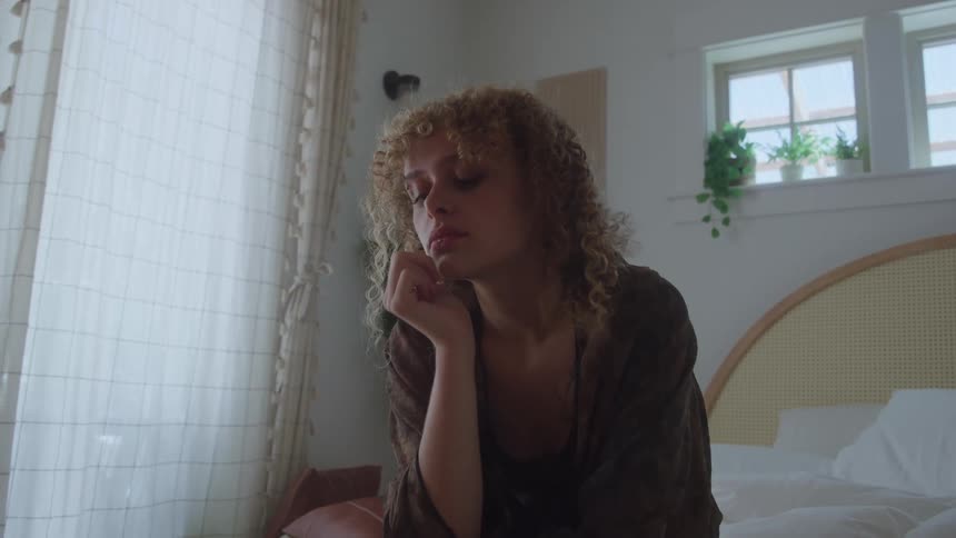 Woman sits on the edge of the bed with her head resting on her hand in a naturally lit residential setting