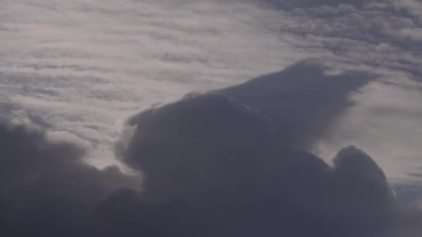 A view from above the clouds shows a massive dark cumulus cloud formation slowly rising and casting a shadow against a layer of bright textured altocumulus clouds in the distance