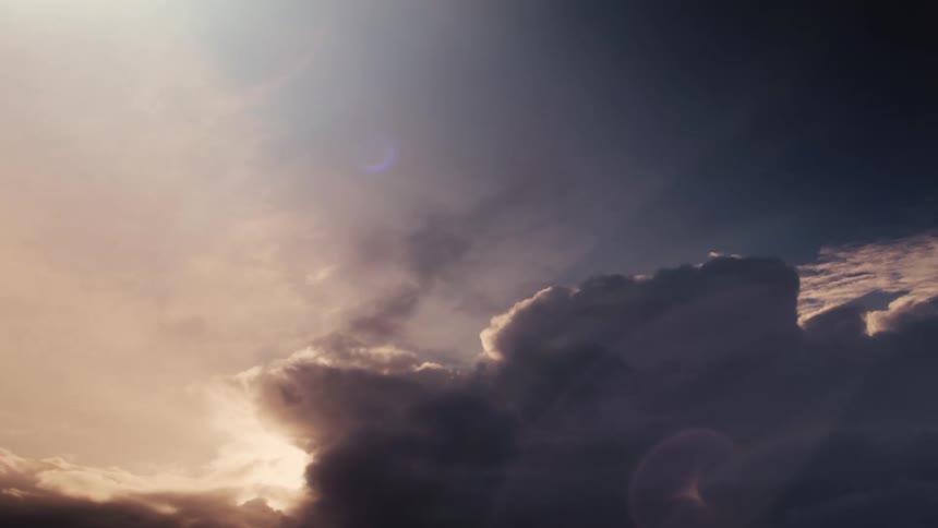A large dramatic dark cloud formation slowly moves across the sky with a bright warm light source from the left casting a vibrant lens flare and illuminating the edges of the clouds