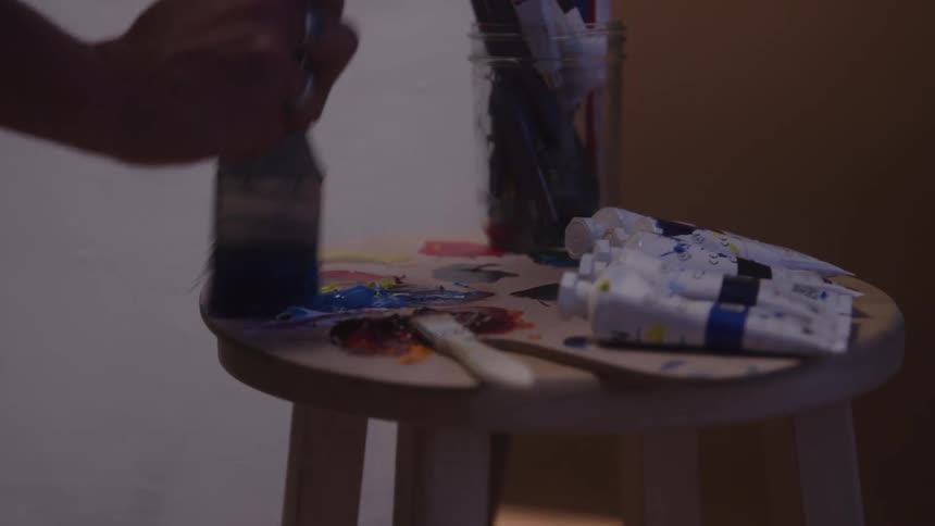 An artists hand dipping a paintbrush into paint on a wooden palette resting on a stool