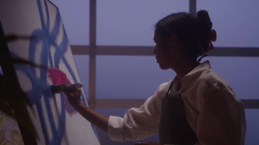 Woman painting abstract blue and pink shapes on a canvas positioned in an indoor setting with soft lighting