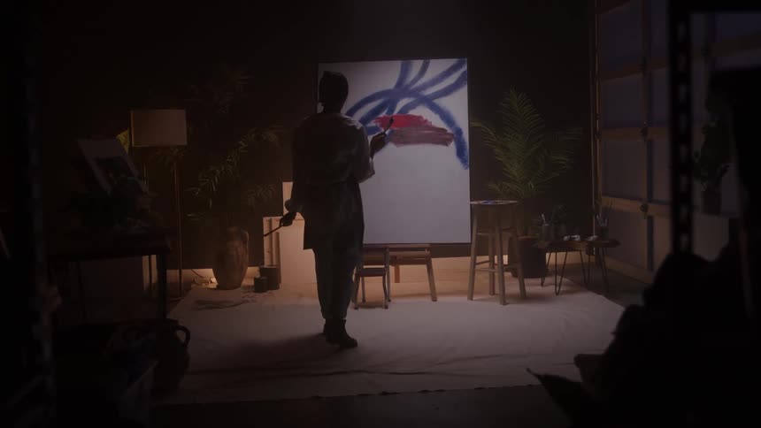 A rear view of an artist in a white shirt standing on a chair painting bold blue and red abstract strokes onto a large white canvas in a dimly lit studio space