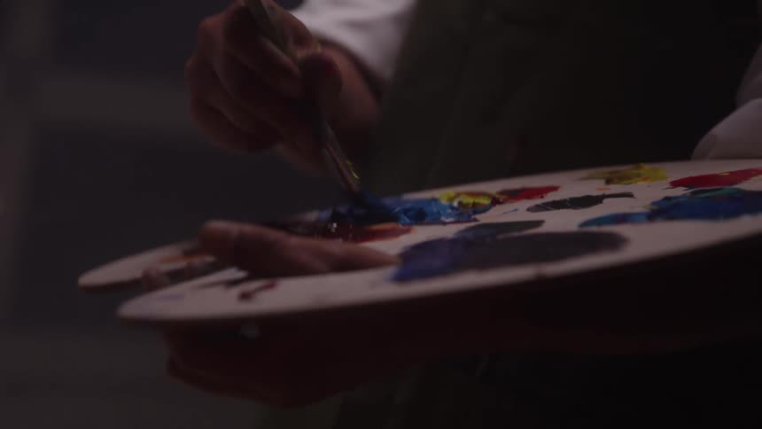 Woman holding a palette with colorful paints while mixing pigments with a brush in an artistic setting with soft lighting