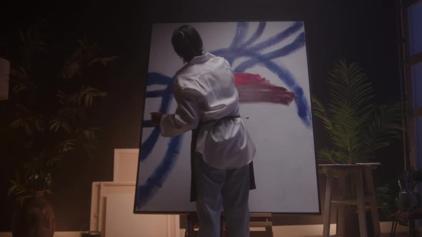 A rear view shows an artist in a white button down shirt and gray pants standing on a stool to paint a large abstract canvas with broad blue and red strokes in a dimly lit studio space with plants and art supplies around