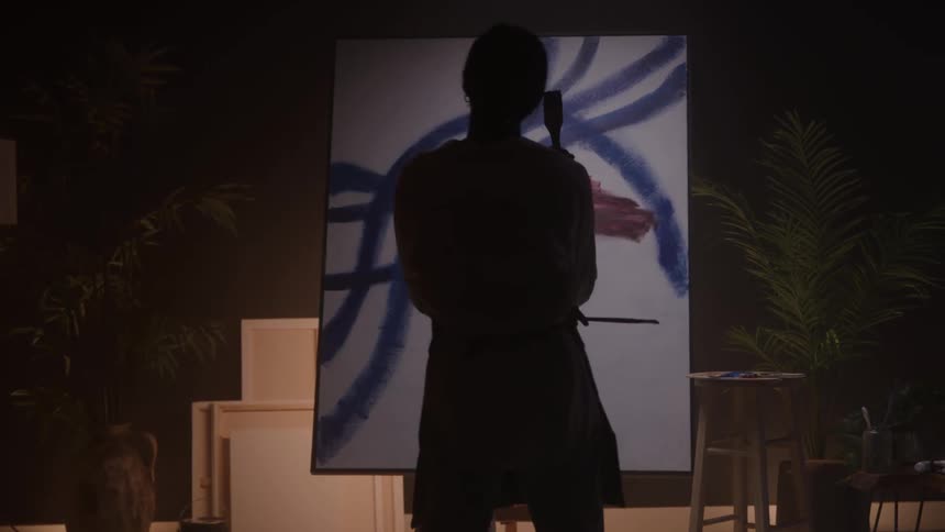 A woman stands in a dark room illuminated by warm light holding a brush as she paints on a canvas