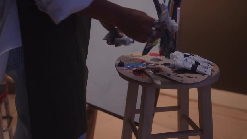 Person preparing paint on a palette beside an easel with painting materials on a wooden stool