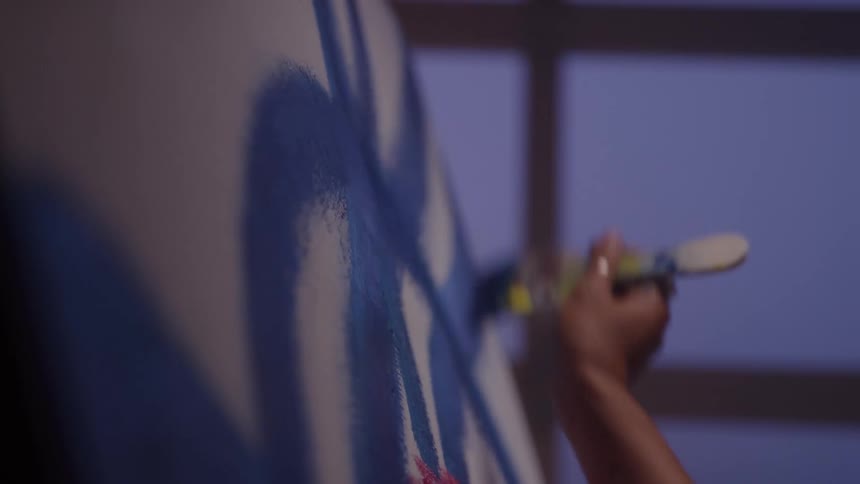 A close view of an artists arm shows a hand holding a paintbrush applying thick blue paint to a white surface against a blurred background of a dark window frame