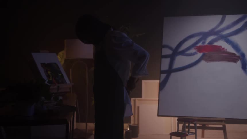 In a dark art studio a silhouetted artist adjusts their apron while standing between two paintings on easels