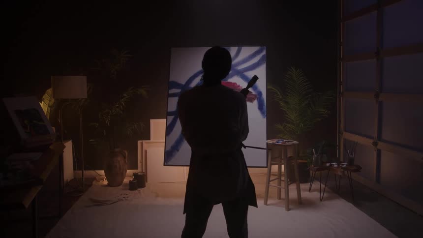 Silhouetted from behind an artist in a dimly lit studio paints with bold strokes on a large spot lit canvas