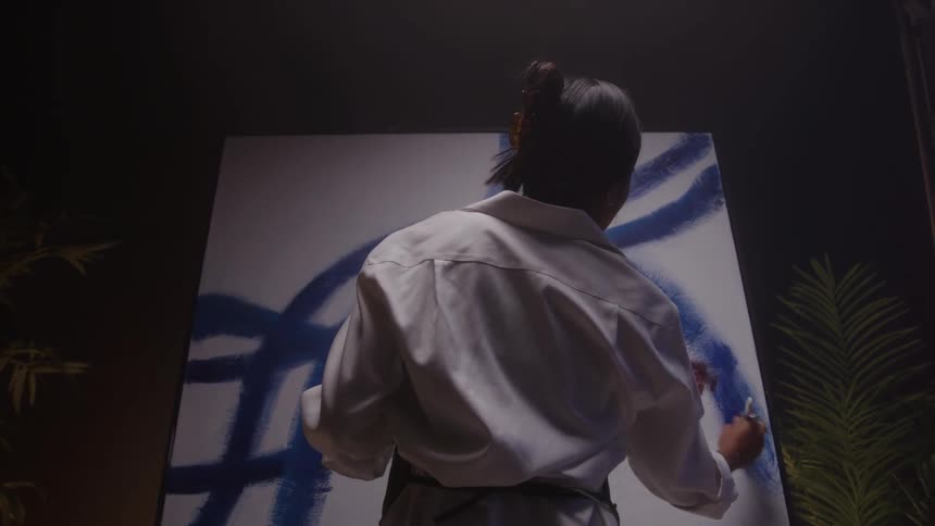 A rear view shows a woman artist in a white shirt and dark pants painting a large canvas with bold blue lines and a splash of red in a dark studio setting
