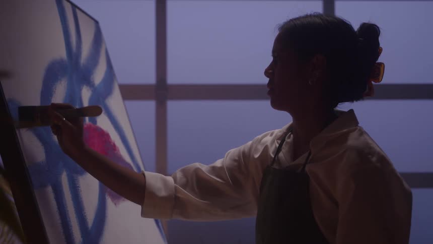 In a dimly lit studio a female artist in an apron is silhouetted as she paints broad strokes onto a large canvas