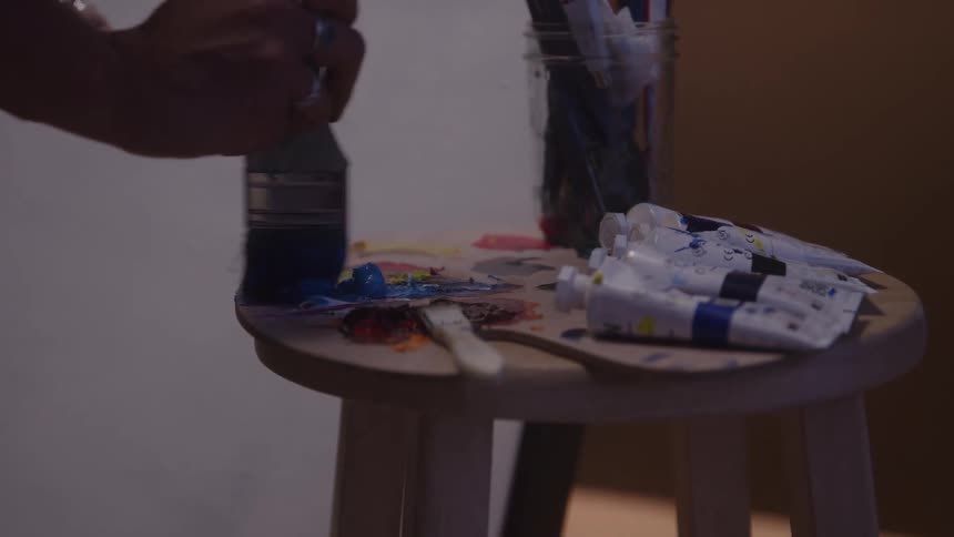 The video shows a closeup of an artist mixing paint on a small wooden palette resting on a stoolseveral tubes of paint are arranged on the stool next to the palette while a hand holding a large paintbrush mixes a dark color