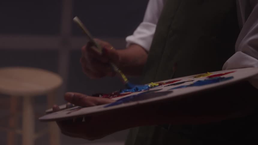 Person holds a paint palette with various colors while mixing paint with a brush in a softly lit indoor setting