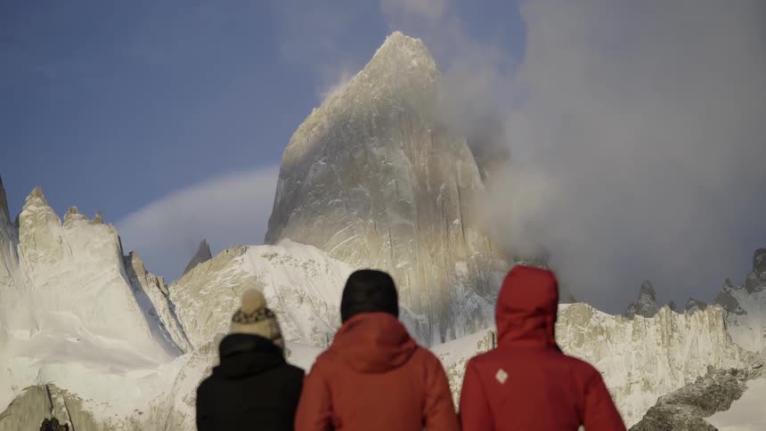 Three people are standing below rocky, snow covered peaks