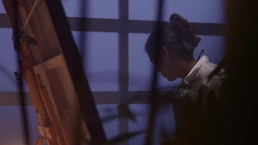 In a dimly lit art studio a female artist leans in closely to her canvas on an easel concentrating on her painting
