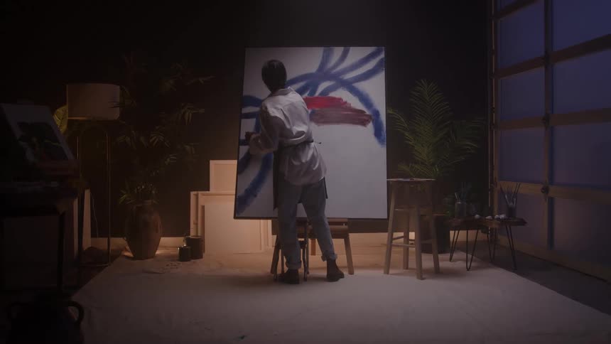 A young female artist paints on a canvas holding a brush in one hand and a paint can in the other applying bold strokes of blue paint with focused intent