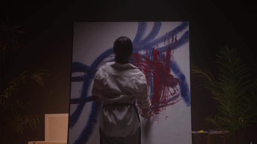 Person paints a canvas with blue lines and red marks in a studio with focused lighting and art supplies nearby