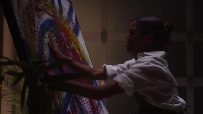 Woman paints an abstract canvas in a lit studio wearing a white shirt and dark apron with tiedback hair
