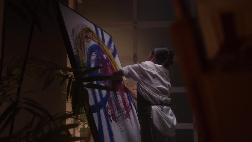 Woman paints a canvas with blue lines and colorful circular shapes in a studio with plants and a garagestyle door in the background