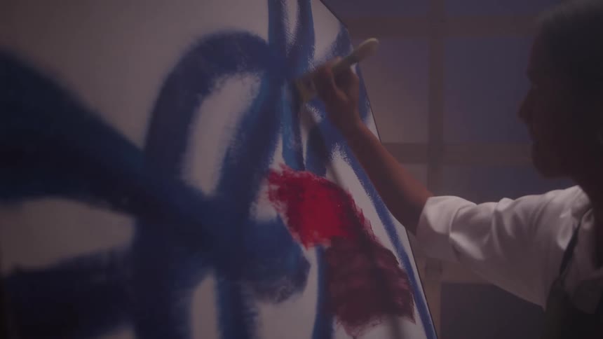 A young woman in a studio carefully strokes a brush loaded with blue paint across a canvas focused intently on crafting a detailed art piece