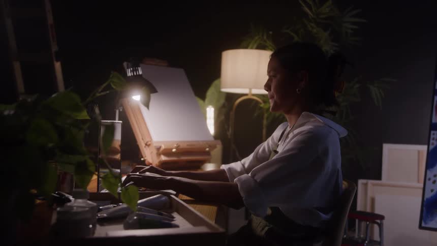Woman sitting in front of a laptop in a creative setting with an easel plants and office supplies on the desk
