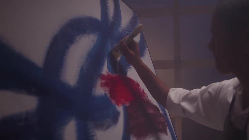 An artist applying bold blue brushstrokes to a large canvas that already has patches of red and blue paint