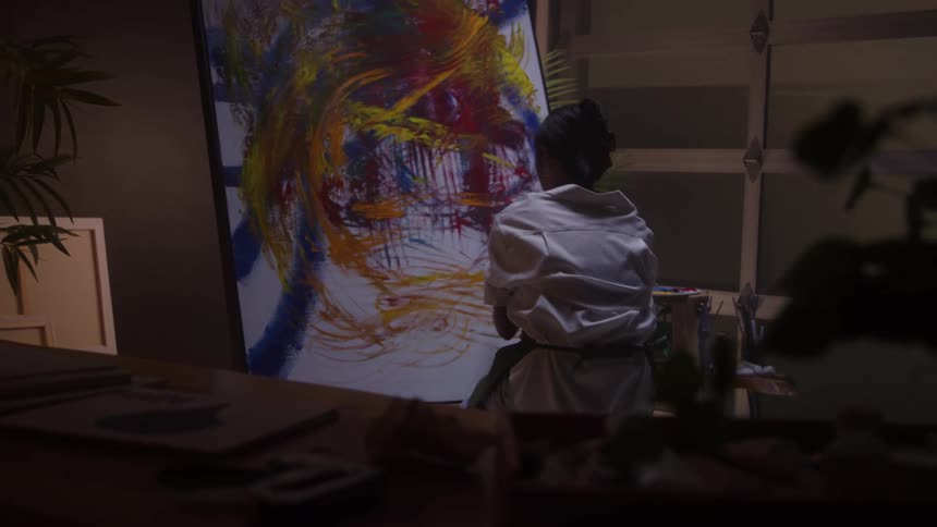 In a dimly lit studio a female artist seen from behind energetically paints a large colorful abstract canvas