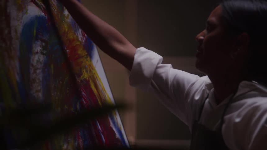 In a dimly lit studio a female artist passionately applies paint with her hand to a colorful abstract canvas