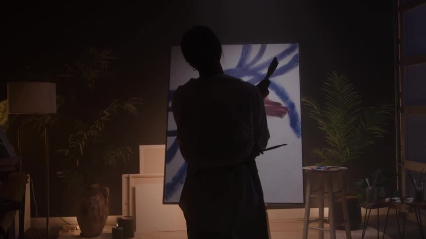 A young black female artist inspecting her canvas painting from a distance in an art studio at night
