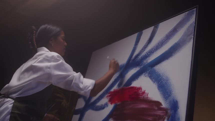 A profile close view shows a woman artist in a white shirt and dark apron painting bold blue lines and red patches onto a large white canvas