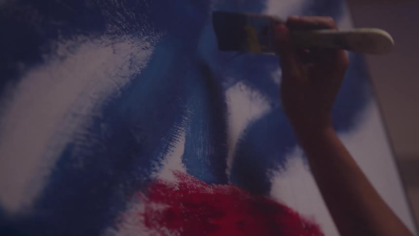 The video shows a closeup of a hand holding a paintbrush moving it across a canvas with bold dark blue paintbeneath the blue patches of bright red paint are visible suggesting the artist is layering colors in an abstract or dynamic style