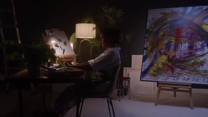 A young black female artist using a laptop computer while sitting at a desk in an art studio at night