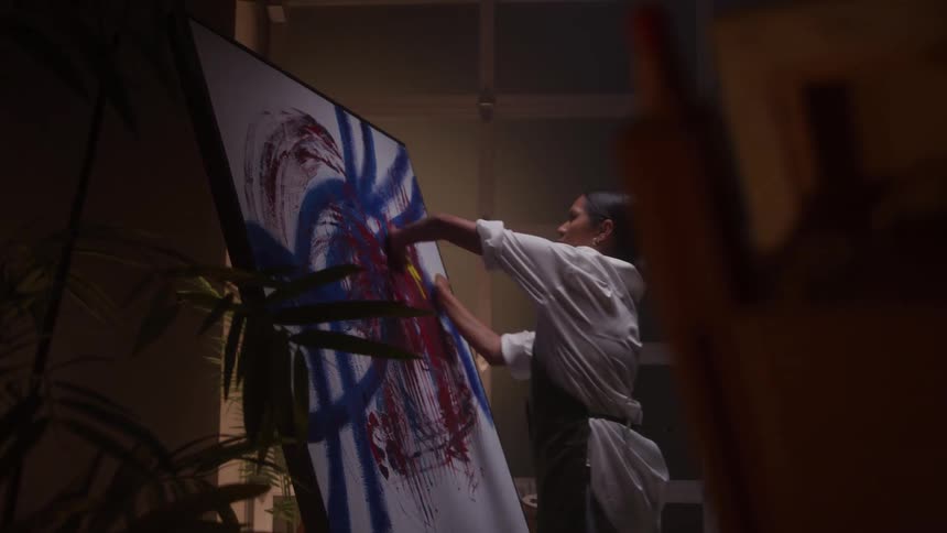 The video shows an artist in an indoor studio space illuminated by a warm table lamp painting at an easel with a large abstract canvas next to her she is dressed in a white buttonup shirt and dark overalls and is seen mixing colors on a small