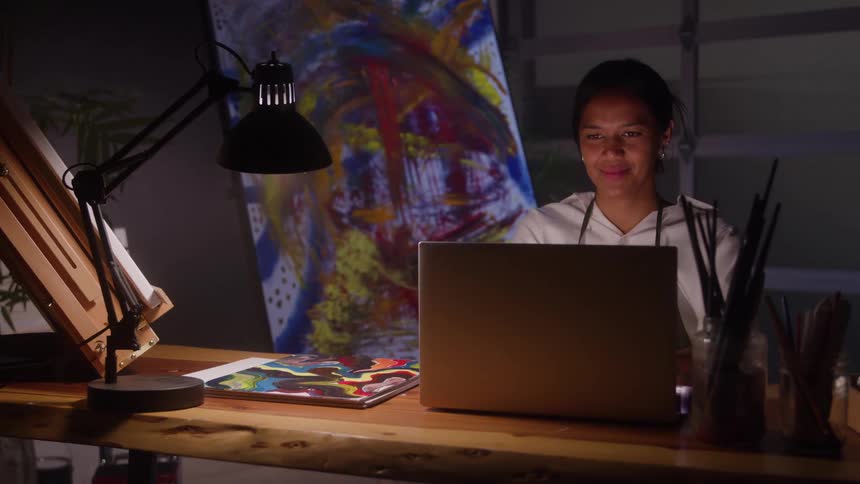 A smiling woman sits at a wooden desk with a lamp and art supplies working on a laptop with a colorful abstract painting visible on an easel in the background of the dark studio
