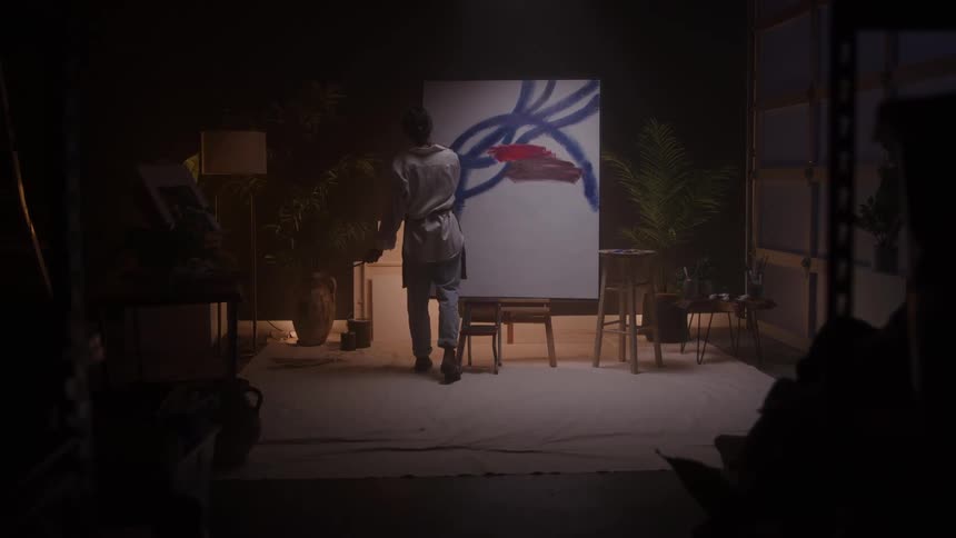 An artist wearing a white outfit stands with her back to the camera painting bold blue and red abstract strokes onto a large white canvas in a dimly lit studio filled with plants