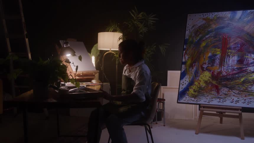 A female artist steps away from her canvas and settles into a chair in her studio working on her laptop