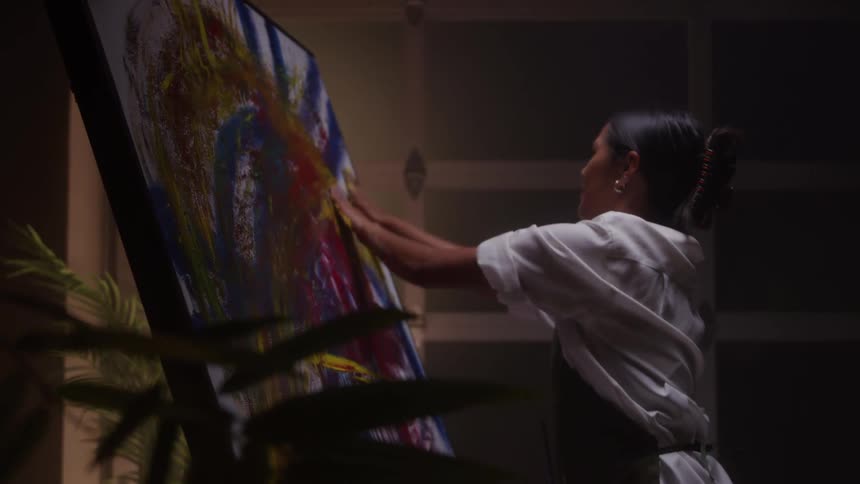 A female artist stands in a dimly lit studio using her palm to paint on a canvas she swings her hand left to right in broad expressive strokes the low light casting soft shadows across the textured surface