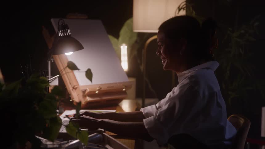 Woman typing on a table illuminated by a lamp in a creative space with plants and an easel