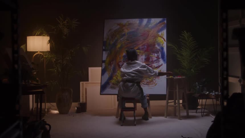 Woman seated in front of the canvas painting vibrant shapes in red yellow blue and brown in a studio with warm lighting and surrounding plants