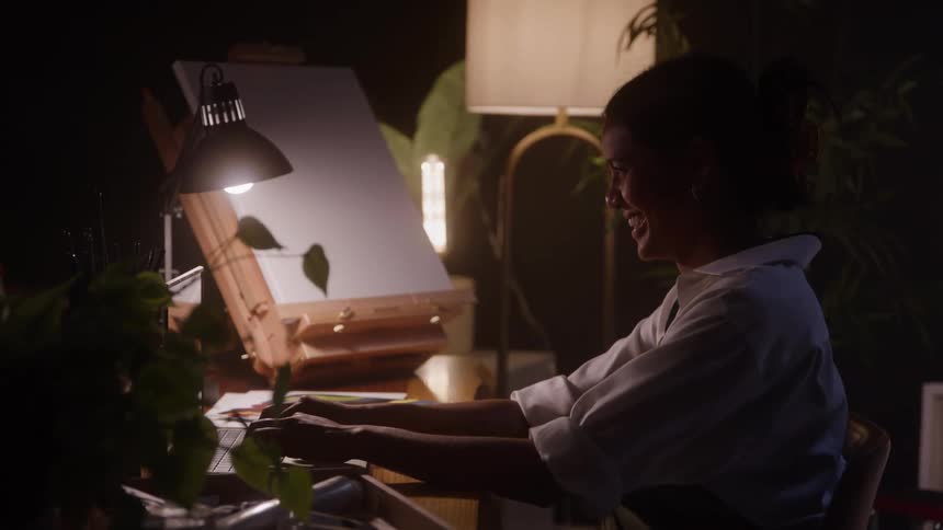 Woman smiles while working on a laptop at a welllit desk with plants and an easel in the background in a creative setting