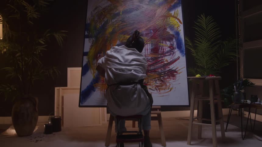 Woman sits on a stool while painting a canvas with colorful brushstrokes in a studio surrounded by plants and art supplies