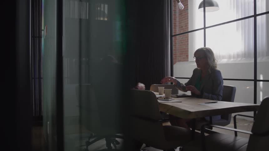 Man and woman shake hands while seated at a table in a modern office with large windows and a glass partition