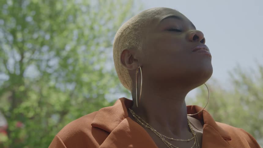 Woman with short blonde hair stands with eyes closed and face tilted upward in an outdoor setting under a blue sky