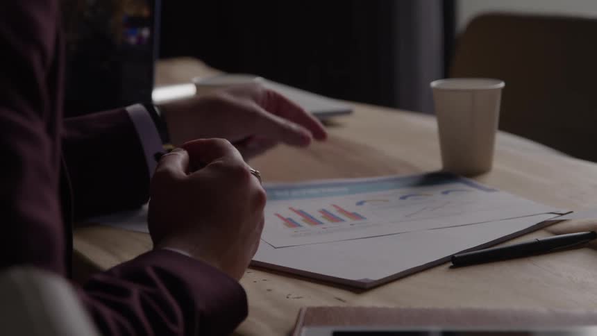A closeup shot shows a person sitting at a table in a dark jacket picking up and looking at a printed document with charts then placing it down and holding a pen with both hands while contemplating
