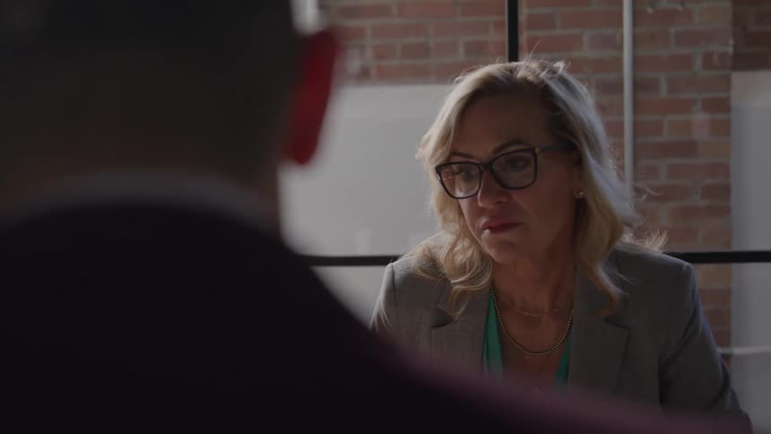 A woman wearing glasses and a suit engages in a discussion with a man indoors both focused and involved in conversation