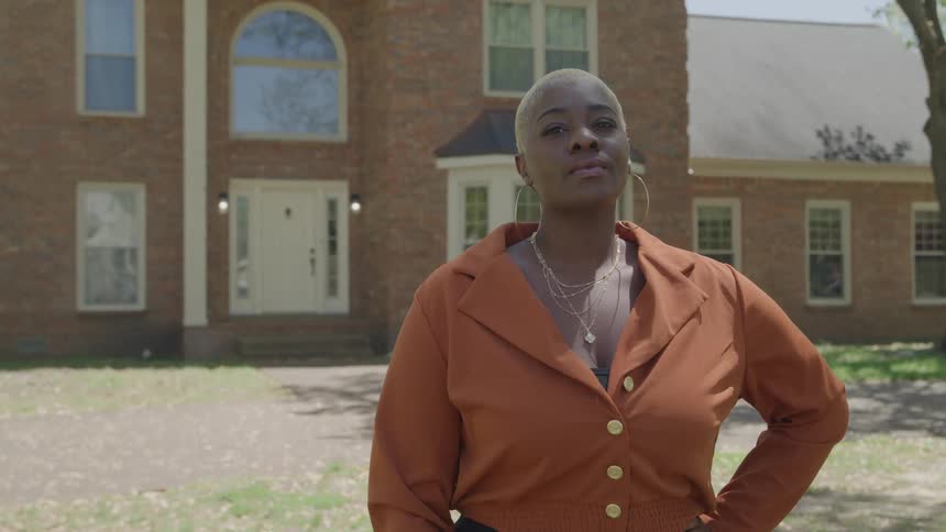 Woman stands in front of a brick house with large windows wearing a buttoned blouse and gold accessories