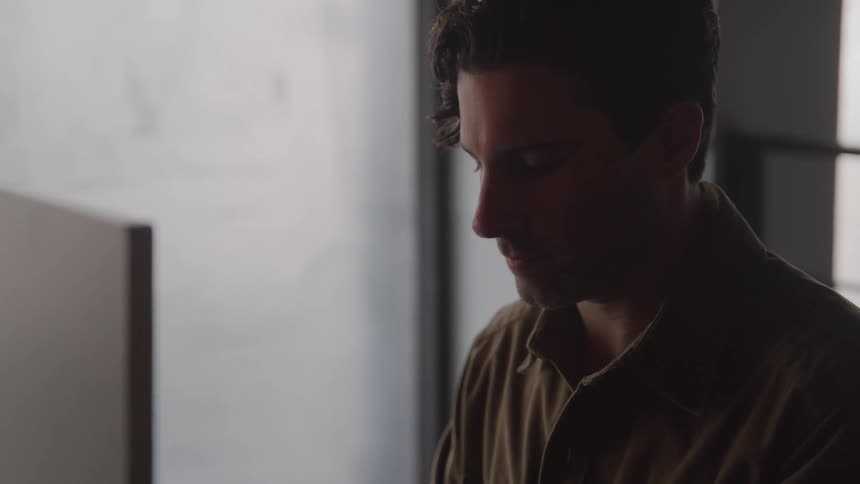 The video is a closeup of a mans hands typing quickly on a silver laptop keyboardhe is wearing a brown or dark collared shirt and a potted plant and coffee cup are visible in the blurry foreground