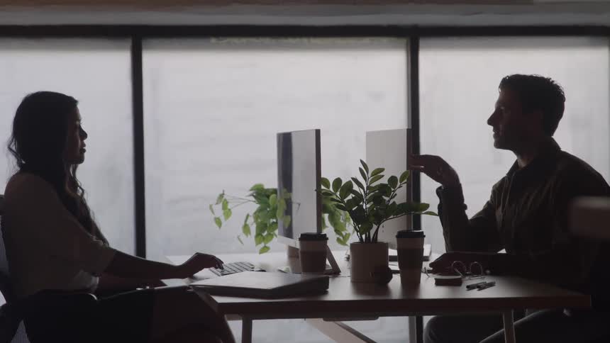 A young woman sits in a cubicle opposite a male coworker both engaged in a midday workhour conversation