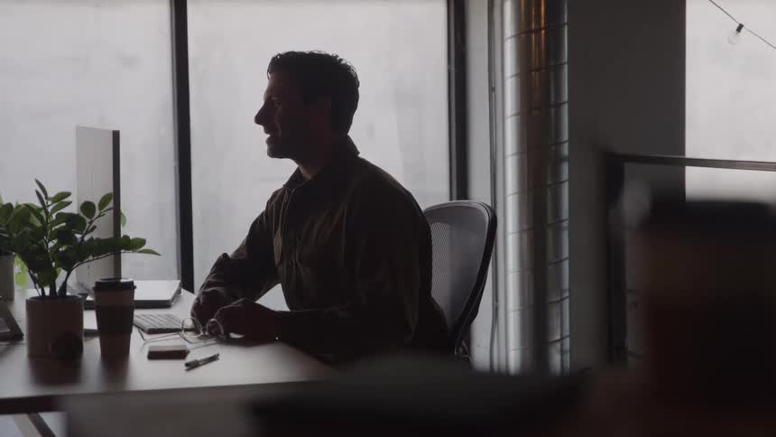 A side view shows a man in a dark green shirt sitting at a desk and writing on paper in a darkly lit office silhouetted against a bright window