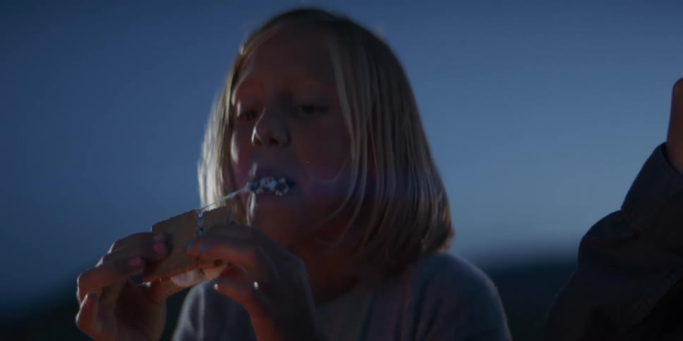 A close view shows a young girl with blonde hair biting into a messy smore sandwich illuminated by the warm light of a campfire against a dark blue evening sky